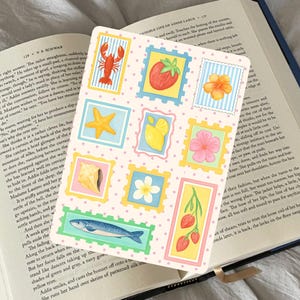 Puede incluir: Una hoja de pegatinas decorativas con ilustraciones coloridas de una langosta, una fresa, una estrella de mar, un limón, una flor y un pez, diseñadas al estilo de un sello postal. La hoja está colocada sobre un libro abierto.
