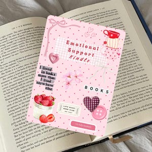 Op de afbeelding: Een roze Kindle-hoes met de tekst "Emotional Support Kindle" en decoratieve elementen, waaronder harten, bloemen en de tekst "I lived in books more than anywhere else."