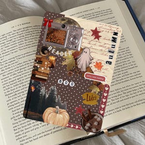 May include: A decorative bookmark with a collage of autumn-themed elements. It features a digital camera, a pumpkin, leaves, a ghost, and the word "Autumn". The bookmark is placed on an open book.