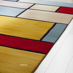 May include: A colorful geometric rug with a modern design. The rug features rectangular blocks in yellow, red, beige, light blue, and gray, outlined with black lines. The rug is placed on a white wooden floor.