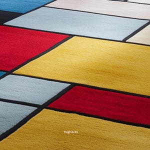 May include: Close-up of a colorful geometric rug with a modern design. The rug features rectangular shapes in red, yellow, blue, and gray, outlined with black borders. The text "RugHacks" is visible in the bottom right corner.