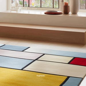 May include: A colorful geometric rug with rectangular blocks in yellow, red, blue, and cream, separated by black lines. The rug has a modern design and is placed on a light-colored wooden floor. The text "RugHacks" is visible in the bottom corner.