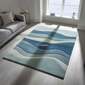Può includere: Un tappeto rettangolare con un motivo a onde nei toni del blu e del bianco. Il tappeto presenta strisce orizzontali di larghezze e colori variabili, creando un aspetto strutturato simile all'oceano. Il testo "Rug Hacks" è visibile sul tappeto.