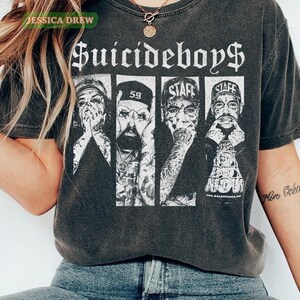 May include: Dark gray t-shirt featuring the band name "Suicideboys$" in a gothic font. The shirt displays four black and white portraits of the band members. The shirt has a vintage, worn look.