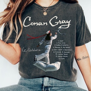 May include: Dark gray t-shirt featuring the text "Conan Gray" in white script above an illustration of a person in a sailor outfit. The shirt also lists song titles from the album "Wishbone".
