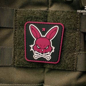 Könnte beinhalten: Ein quadratischer Aufnäher mit einem rosa Kaninchenkopf mit wütendem Gesichtsausdruck und gekreuzten Knochen auf schwarzem Hintergrund. Der Aufnäher hat einen rosa Rand und befindet sich auf einem grünen taktischen Hintergrund. Der Text "PREMIUM PATCHES BY SEVAL" steht oben rechts.