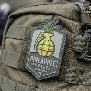 Könnte beinhalten: Ein rechteckiger Aufnäher mit einem Ananas-Granaten-Design, mit gelben und grünen Farben und dem Text "PINEAPPLE EXPRESS AIRMAIL DELIVERY". Der Aufnäher ist an einem grünen Rucksack befestigt.