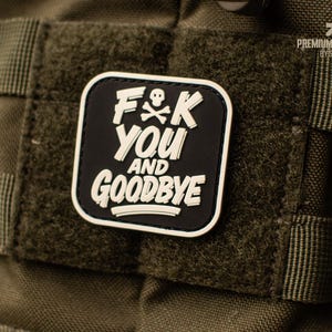 Könnte beinhalten: Ein schwarz-weißer Aufnäher mit dem Text "F*K YOU and GOODBYE" und einem Totenkopf- und gekreuzten Knochen-Symbol. Der Aufnäher ist quadratisch mit abgerundeten Ecken und an einem grünen Stoff befestigt.