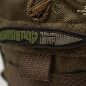 Könnte beinhalten: Ein schwarzer und grüner Aufnäher mit den Worten "EVERYDAY COMPANION" in einem messerförmigen Design. Der Aufnäher befindet sich auf einer strukturierten grünen Oberfläche. Der Aufnäher trägt auch den Text "PREMIUM PATCHES BY SEVAL".