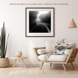 May include: Black and white framed print of a road disappearing into a foggy forest. The print is displayed in a living room setting with a wooden chair, decorative pillows, and a potted plant. The text at the top says "SHOWN FRAMED AS EXAMPLE ONLY - PRINTS COME UNFRAMED."