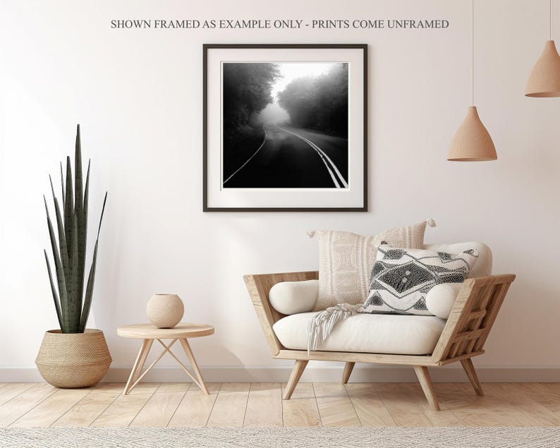 May include: Black and white framed print of a road disappearing into a foggy forest. The print is displayed in a living room setting with a wooden chair, decorative pillows, and a potted plant. The text at the top says "SHOWN FRAMED AS EXAMPLE ONLY - PRINTS COME UNFRAMED."