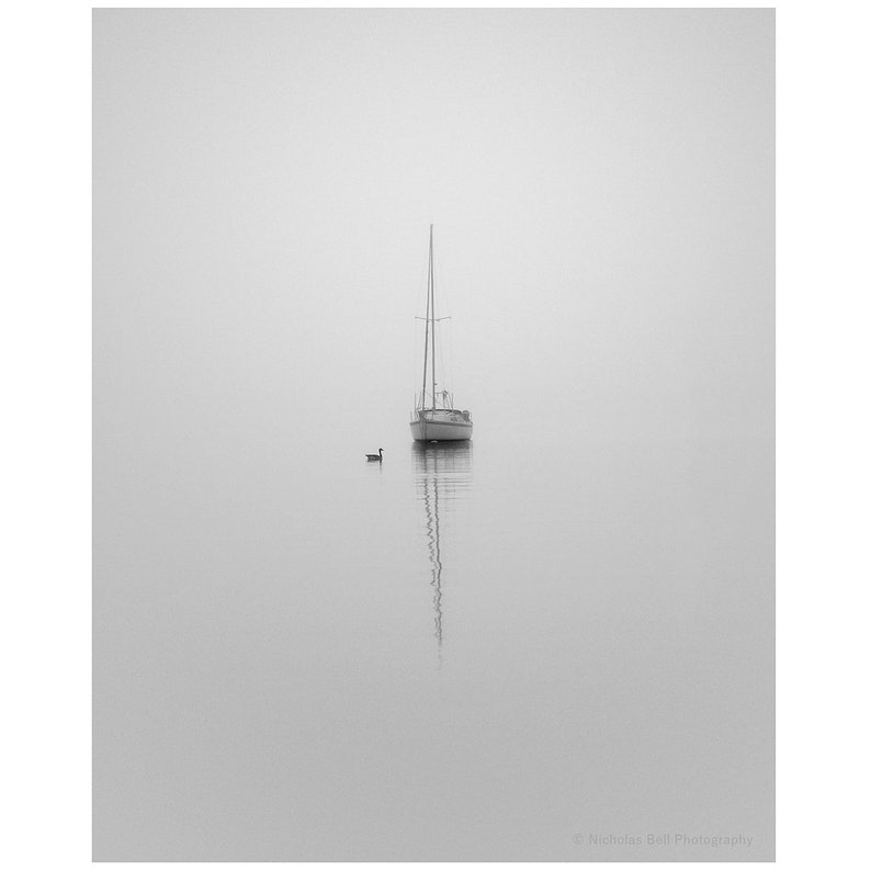 Op de afbeelding: Een zeilboot ligt voor anker in een stil water op een mistige dag. Een enkele eend zwemt in de verte.