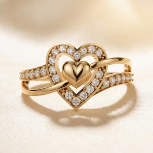 May include: A gold-colored ring featuring a heart-shaped design. The ring has a central heart shape surrounded by a halo of small, clear gemstones. Additional gemstones are set along the band, creating a sparkling effect. The ring is set against a soft, off-white background.