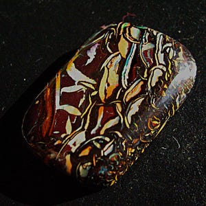 May include: A polished, rectangular gemstone with a complex pattern of colors. The stone features a dark maroon base with veins of white, yellow, and orange. The surface is smooth and reflective, with a slight curve.