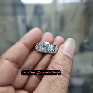 May include: A silver ring with a large, round, clear gemstone at the center. The band features decorative cutouts. The ring is held in a hand. The text "Aradhya Jeweller Shop" is at the bottom.