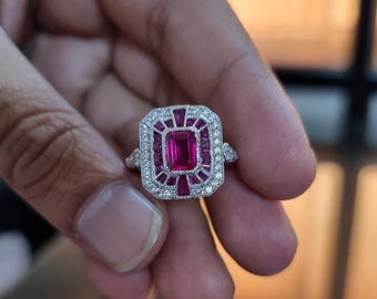 May include: A silver ring with a rectangular, vibrant pink center stone, surrounded by smaller pink and white stones. The ring has a geometric design, with a double halo of white stones. The ring is held up by a person's hand.