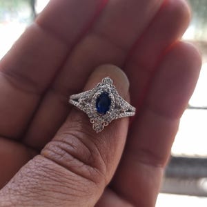 May include: A silver ring featuring a large, oval, dark blue gemstone. The ring has an intricate filigree design, with smaller, clear gemstones encircling the central stone. The ring is held on a person's finger.