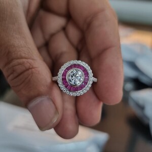 May include: A silver ring featuring a central clear gemstone, encircled by a band of small, rectangular, red gemstones. The outer ring is adorned with small, clear gemstones. The ring is held in a hand.