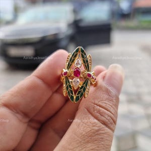 May include: Gold-toned ring featuring a central oval design with a ruby gemstone, flanked by white and red gemstones. The ring has green enamel accents and is held in a hand. The text "Aaradhya JewelShop" is visible.