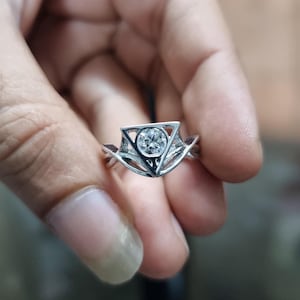 May include: A silver ring with a central, round, clear gemstone. The ring features a geometric design with a triangle shape around the gemstone and interwoven bands. The ring is held in a hand.