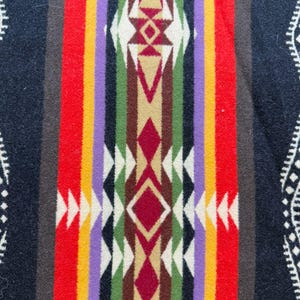 May include: A woven textile featuring a central vertical band with geometric diamond and triangle patterns in red, green, brown, purple, yellow, and white. The background is dark navy blue with white decorative border accents on the outer edges.