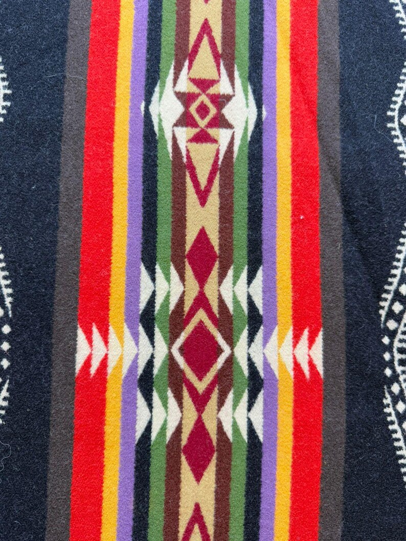 May include: A woven textile featuring a central vertical band with geometric diamond and triangle patterns in red, green, brown, purple, yellow, and white. The background is dark navy blue with white decorative border accents on the outer edges.