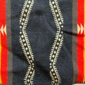 May include: A woven blanket featuring a central black panel with a cream-coloured diamond wave pattern. The design is flanked by vertical stripes in brown, vibrant red, and yellow, with small cream-coloured arrow motifs on the red sections.