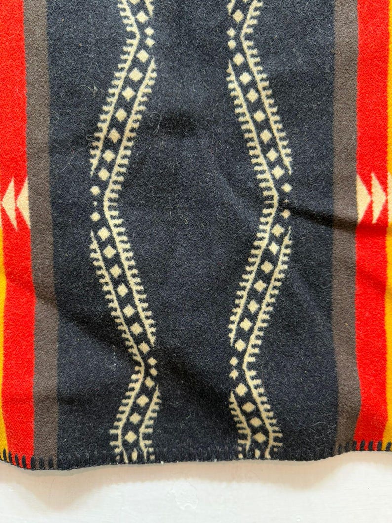 May include: A woven blanket featuring a central black panel with a cream-coloured diamond wave pattern. The design is flanked by vertical stripes in brown, vibrant red, and yellow, with small cream-coloured arrow motifs on the red sections.