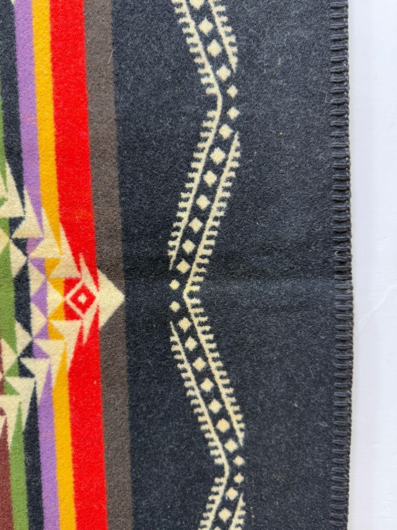 May include: A dark charcoal grey blanket featuring a vertical pattern of vibrant stripes in red, yellow, purple, and green, alongside a geometric diamond-shaped motif. The edge is finished with a dark grey whipstitch border detail.