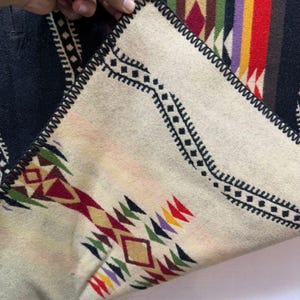 May include: A folded textile featuring a cream-coloured side with black geometric patterns and multi-coloured diamond motifs in red, yellow, green, and purple. The reverse side displays a black background with vertical stripes in red, yellow, and purple.