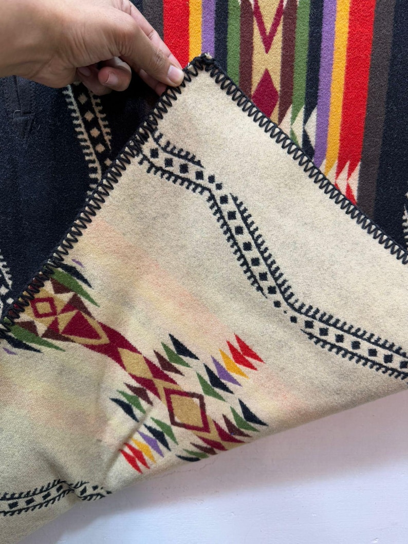 May include: A folded textile featuring a cream-coloured side with black geometric patterns and multi-coloured diamond motifs in red, yellow, green, and purple. The reverse side displays a black background with vertical stripes in red, yellow, and purple.