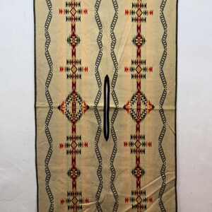May include: A rectangular beige poncho featuring a central vertical slit for the head. The garment displays symmetrical geometric patterns in red, black, green, and yellow, with wavy black lines and a black stitched border along the edges.