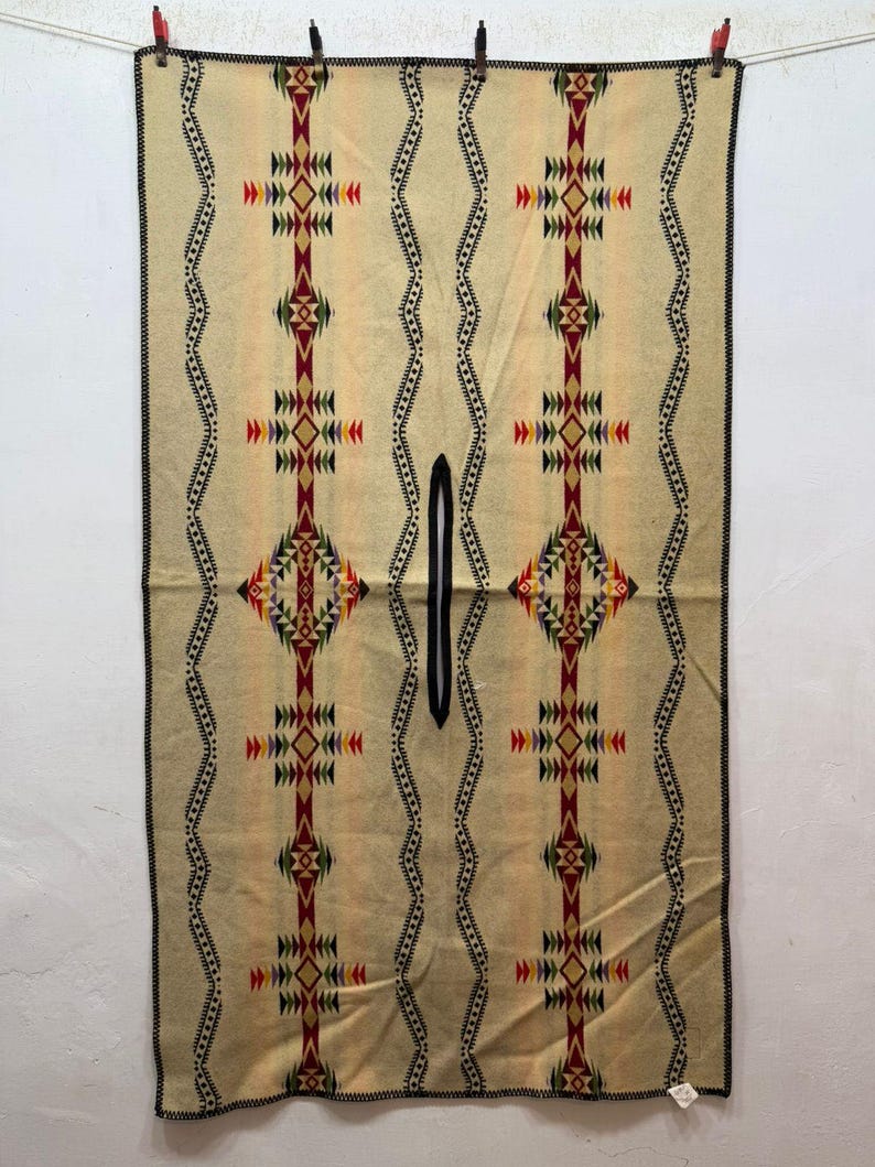 May include: A rectangular beige poncho featuring a central vertical slit for the head. The garment displays symmetrical geometric patterns in red, black, green, and yellow, with wavy black lines and a black stitched border along the edges.