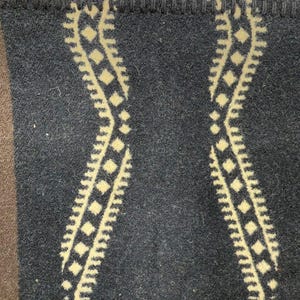 May include: A dark charcoal grey wool blanket featuring a repeating cream-coloured wavy geometric pattern with diamond shapes. The top edge is finished with a thick, dark grey blanket stitch border, and a solid brown section is visible on the left.