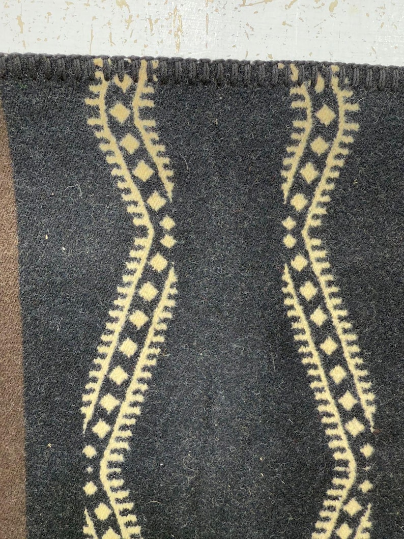 May include: A dark charcoal grey wool blanket featuring a repeating cream-coloured wavy geometric pattern with diamond shapes. The top edge is finished with a thick, dark grey blanket stitch border, and a solid brown section is visible on the left.