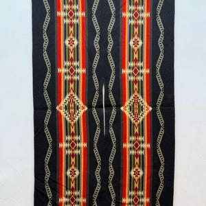 Op de afbeelding: Een zwarte, rechthoekige poncho met een centrale verticale opening voor het hoofd. Het kledingstuk toont symmetrische, kleurrijke geometrische patronen in rood, geel, groen, paars en crème, geaccentueerd door dunne, golvende witte lijnen.