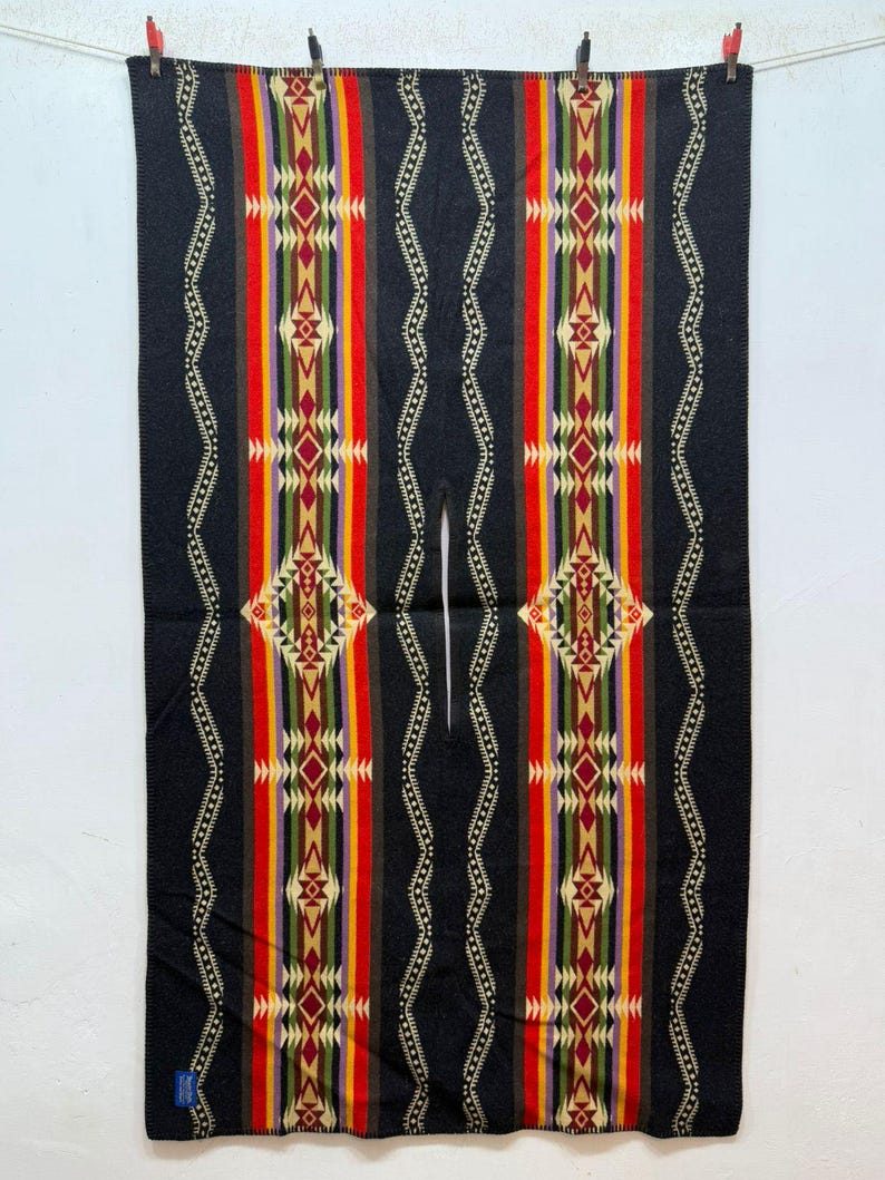 May include: A black rectangular poncho featuring a central vertical slit for the head. The garment displays symmetrical, colourful geometric patterns in red, yellow, green, purple, and cream, accented by thin, wavy white lines on both sides.
