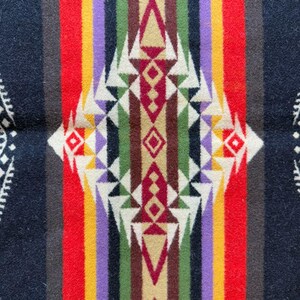May include: A woven textile blanket featuring a symmetrical geometric pattern with a central diamond motif. The design incorporates vertical stripes in black, red, yellow, purple, green, and brown, accented by white triangular shapes.