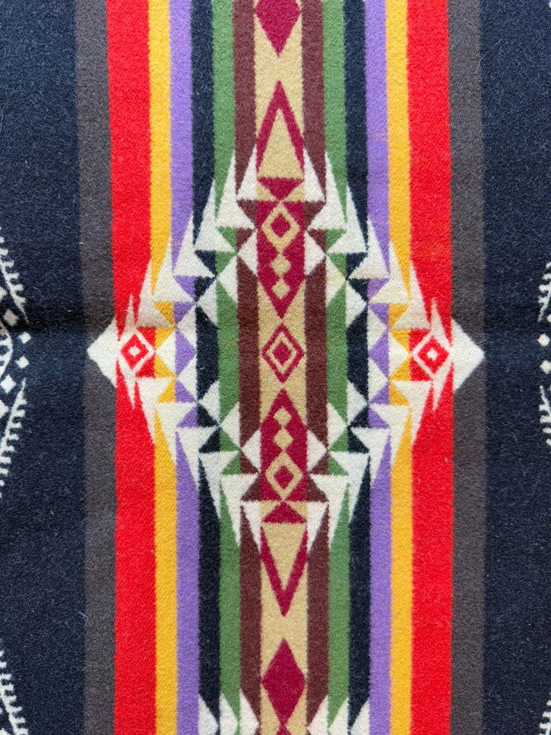 May include: A woven textile blanket featuring a symmetrical geometric pattern with a central diamond motif. The design incorporates vertical stripes in black, red, yellow, purple, green, and brown, accented by white triangular shapes.