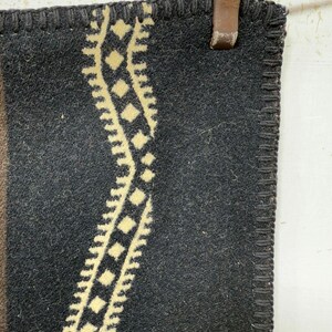 May include: A dark charcoal grey wool blanket featuring a thick, cream-coloured vertical stripe with a repeating diamond and zigzag pattern. The edges of the blanket are finished with a dark, heavy-duty blanket stitch along the entire perimeter.
