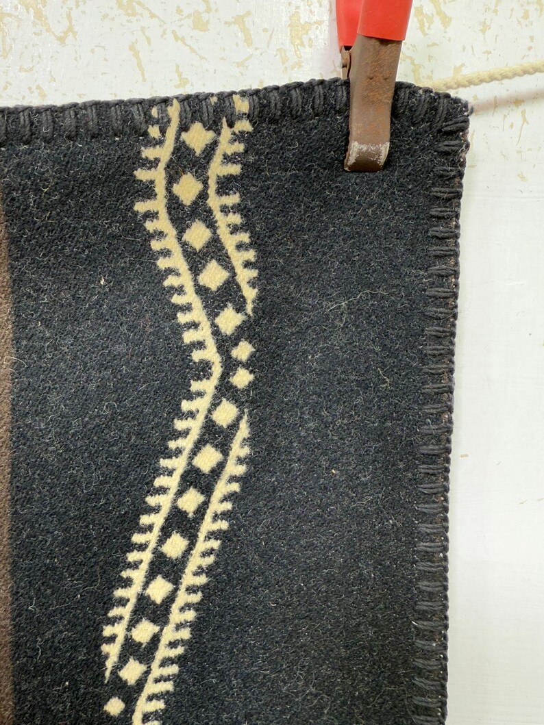 May include: A dark charcoal grey wool blanket featuring a thick, cream-coloured vertical stripe with a repeating diamond and zigzag pattern. The edges of the blanket are finished with a dark, heavy-duty blanket stitch along the entire perimeter.