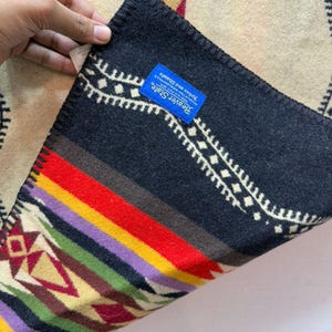 May include: A folded wool blanket featuring a geometric pattern in black, red, yellow, green, and purple. A blue rectangular tag is sewn onto the black section with the text Beaver State, Pendleton Woolen Mills, Robes and Shawls printed in white.