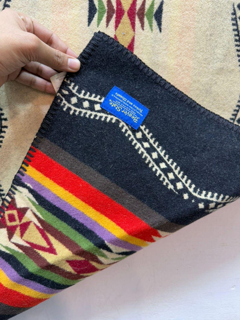 May include: A folded wool blanket featuring a geometric pattern in black, red, yellow, green, and purple. A blue rectangular tag is sewn onto the black section with the text Beaver State, Pendleton Woolen Mills, Robes and Shawls printed in white.