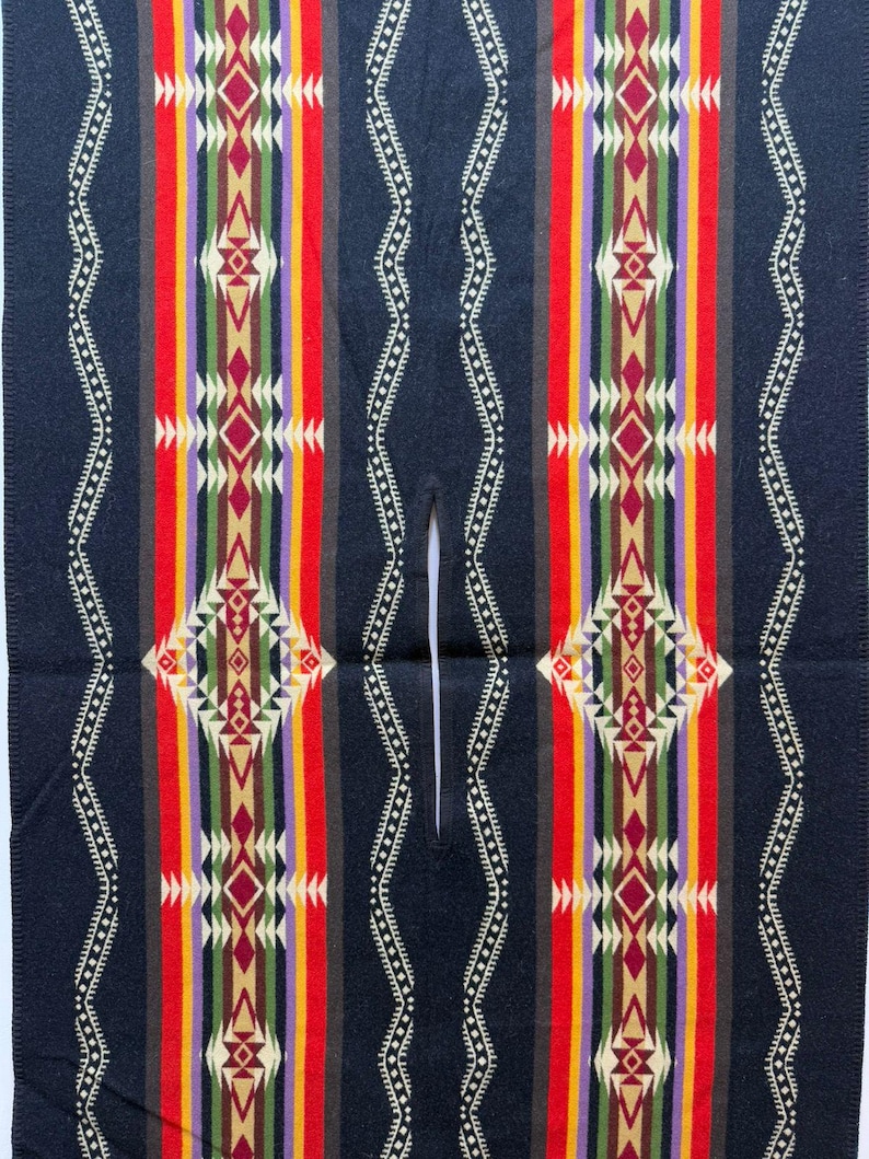 May include: A dark navy blue poncho featuring a central vertical slit opening. The garment is decorated with symmetrical, colourful geometric patterns in red, yellow, green, purple, and cream, arranged in repeating vertical stripes and diamond shapes.