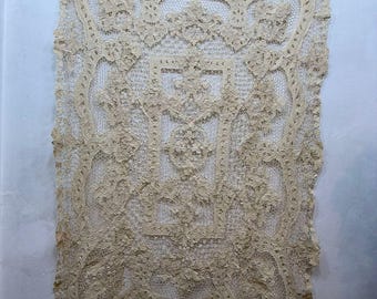 May include: A beige lace textile, possibly a doily or decorative piece, hangs against a white wall. The lace features an intricate floral pattern with a scalloped edge. It is held in place by several clothes pegs.