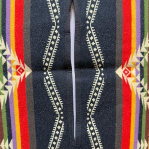 May include: A poncho featuring a central vertical slit opening with black stitched edges. The fabric displays a symmetrical pattern of wide vertical stripes in red, yellow, purple, and green, accented by cream-coloured geometric diamond motifs.