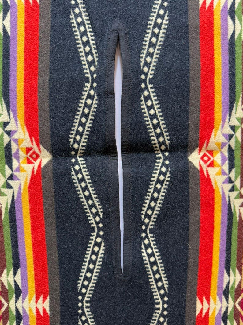 May include: A poncho featuring a central vertical slit opening with black stitched edges. The fabric displays a symmetrical pattern of wide vertical stripes in red, yellow, purple, and green, accented by cream-coloured geometric diamond motifs.