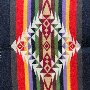 May include: A woven textile featuring a symmetrical geometric pattern with a central diamond motif. The design incorporates vertical stripes in black, red, yellow, purple, green, and brown, accented by white triangular shapes throughout the composition.