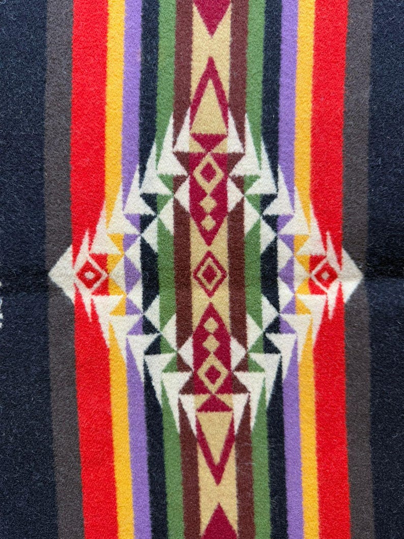 May include: A woven textile featuring a symmetrical geometric pattern with a central diamond motif. The design incorporates vertical stripes in black, red, yellow, purple, green, and brown, accented by white triangular shapes throughout the composition.