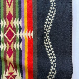 May include: A woven blanket featuring a geometric pattern with vertical stripes in red, yellow, purple, green, and cream. The right side displays a large black section with a cream-coloured wavy line pattern and a finished stitched edge.
